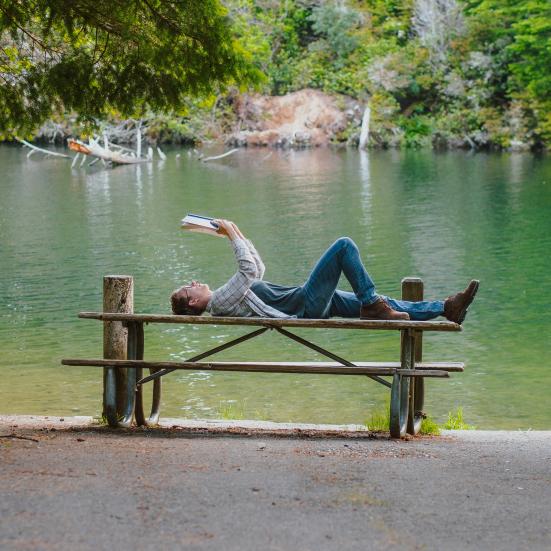 mand der ligger på bord og læser foran en skovsø