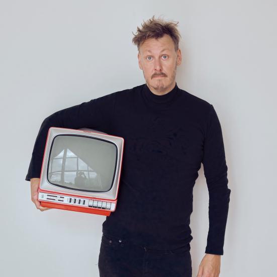 Foto af forsker Andreas Lieberoth i sort tøj med et lille rødt gammeldags fjernsyn under armen, grå baggrund