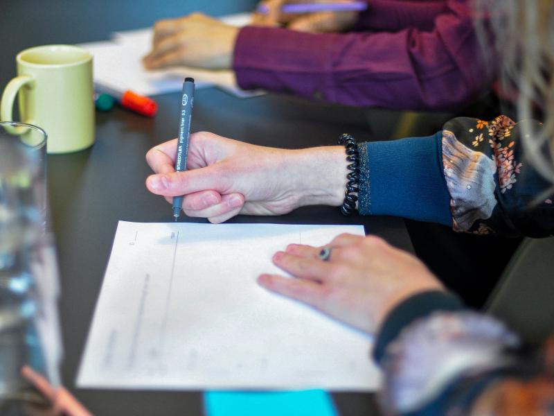 foto af bord med udsnit af mennesker, der skriver på papir. omgivet af glas og krus