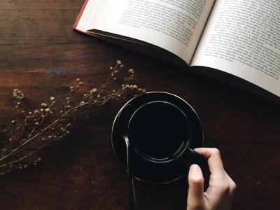 En bog der ligger opslået på et bord og en hånd der holder en kop kaffe