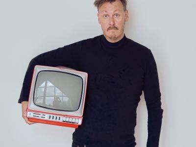 Foto af forsker Andreas Lieberoth i sort tøj med et lille rødt gammeldags fjernsyn under armen, grå baggrund